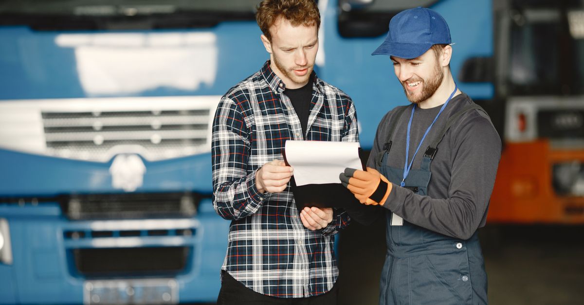 Two men in discussion over vehicle maintenance documents in a garage setting with trucks in the background.
